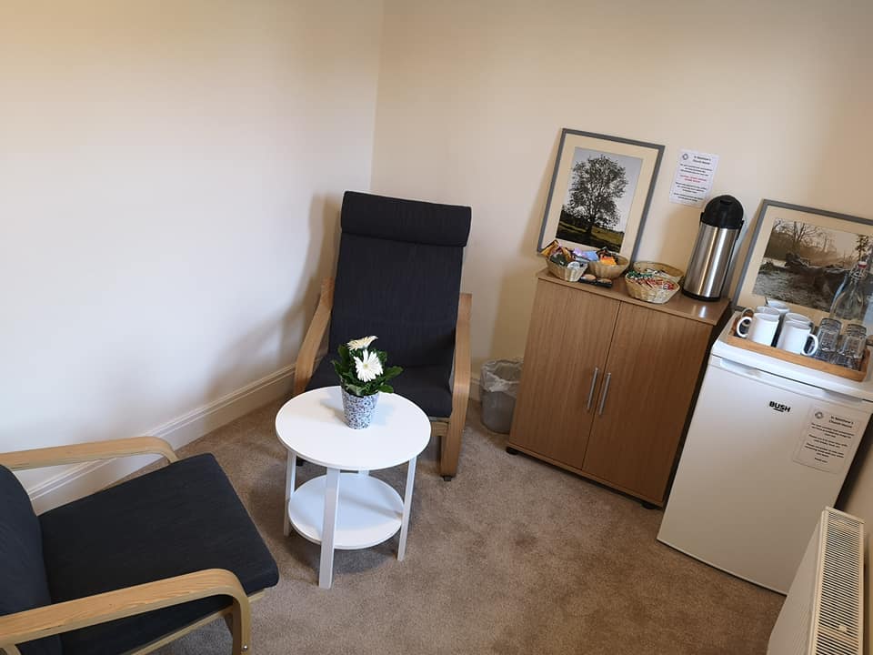 image of St Werburgh's room showing two comfortable chairs facing each other, with a small round coffee table inbetween. In the corner is a fridge and cupboard on top of which is  tea and coffee making area